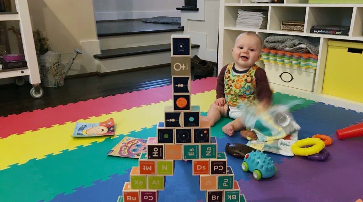 A baby sits on a floor mat smiling.