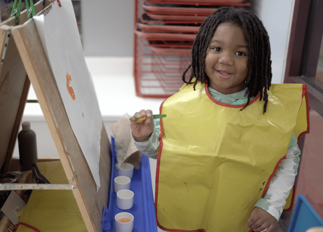 A young child with long hair wearing a yellow smock smiles while holding a paintbrush at an easel, with cups of paint and a partially painted paper in front of them.