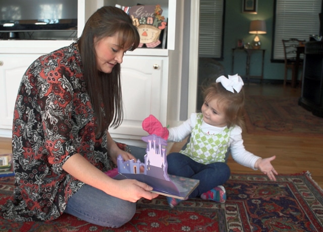 A woman and a young girl sit on a patterned rug, reading a pop-up book together. The girl, wearing a white bow and green vest, gestures with her hands while the woman holds the open book.