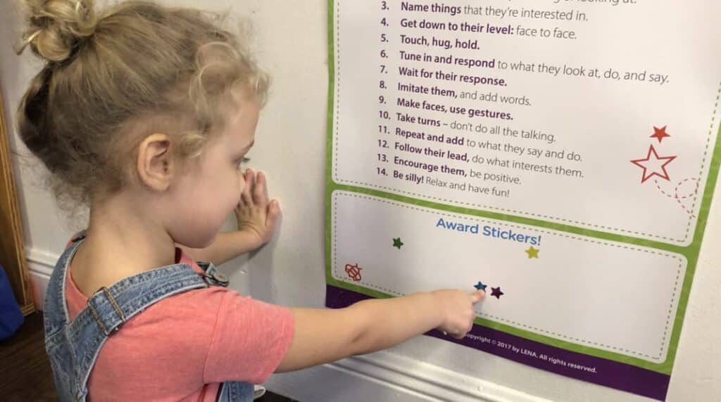 A young child in denim overalls points to colorful star stickers on a Lena Grow wall chart that lists helpful tips and award stickers in a classroom or learning environment.