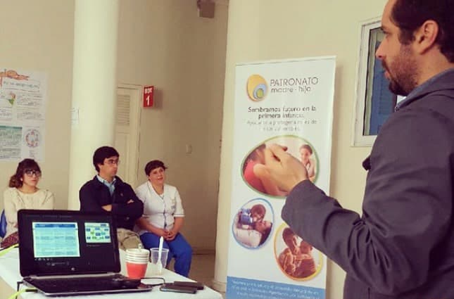 A man gives a presentation to three seated people in a small room. A laptop and disposable cups are on the table, and a standing banner promoting early childhood care and socioemotional development is visible in the background.