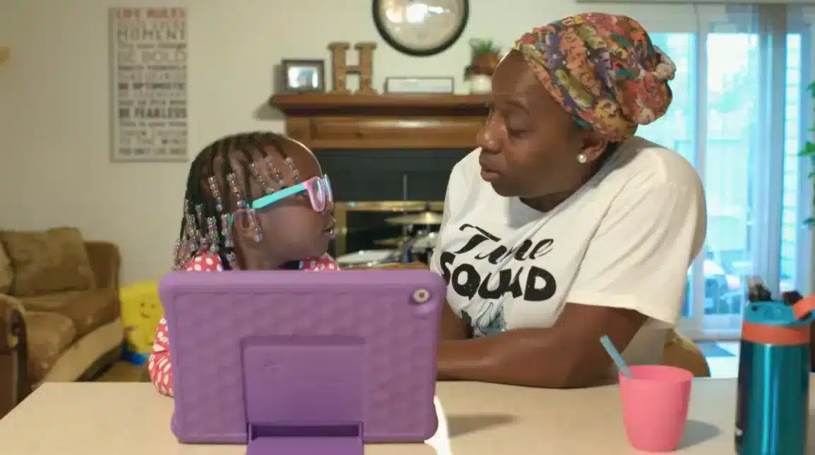 A young girl with beaded hair and glasses sits at a table facing an older woman in a headscarf. They look at each other seriously, with a purple tablet, cup, and water bottle on the table in front of them.