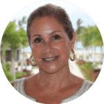 Smiling woman with light brown hair pulled back, wearing gold hoop earrings and a white top, standing outdoors with palm trees and a pathway in the background.
