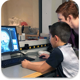 A child and an adult look at a computer screen displaying a brain scan. The adult is seated next to the child, pointing at the screen. The room contains scientific equipment and anatomical models are visible in the background.