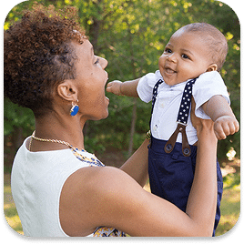A woman joyfully holds a smiling baby boy in her arms outdoors. She wears a white top and earrings, while the baby is dressed in a white shirt with dark suspenders. Trees and greenery are in the background.