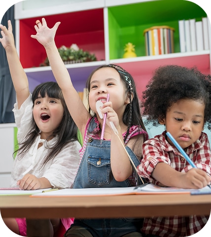 Three children are sitting at a table. The two on the left are raising their hands enthusiastically, while the child on the right is focused on writing. Colorful shelves are in the background.
