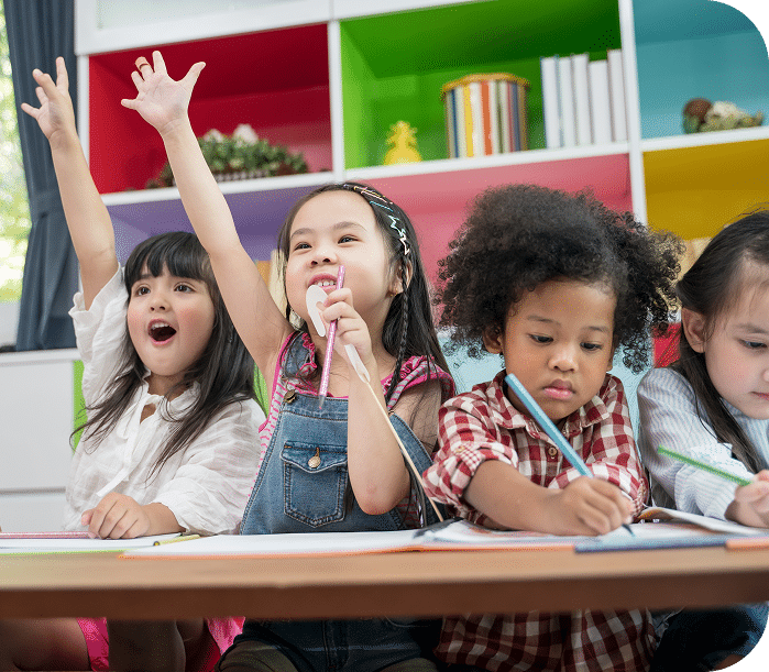 Four young children sit at a table in a colorful classroom. Two girls excitedly raise their hands, while the other two children focus on writing or drawing in their notebooks.