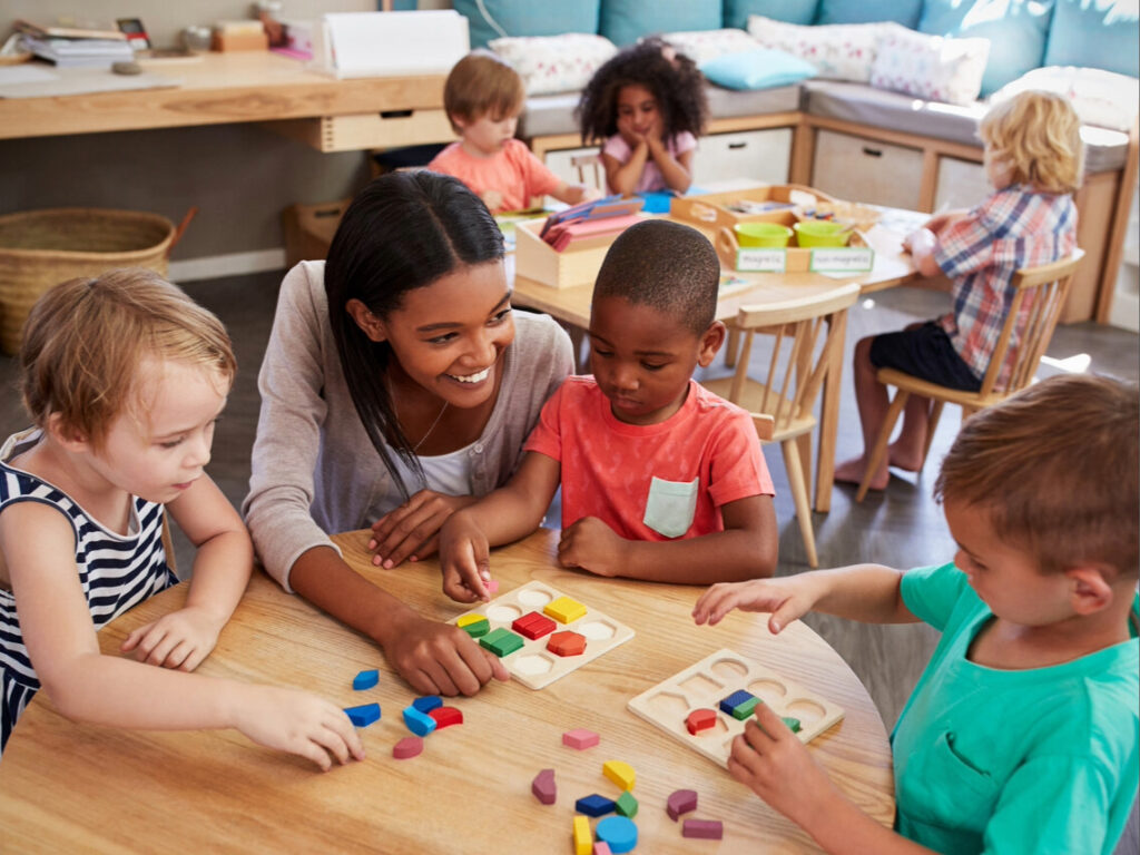 A teacher sits at a table with three young children, helping them play with colorful geometric blocks. In the background, other children play and read in a bright classroom.