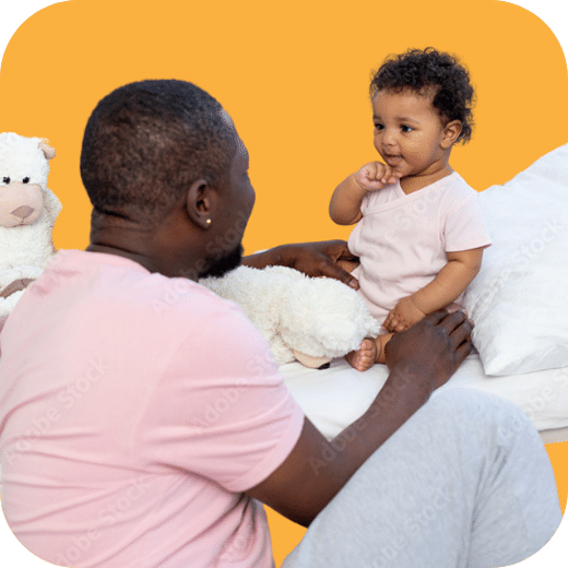 A smiling man in a pink shirt plays with a baby sitting on a bed, holding a white stuffed animal. The baby faces him, and both appear happy, against a solid blue background.
