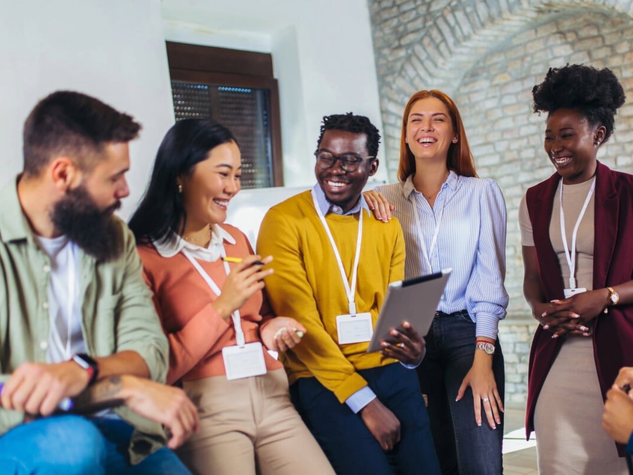 Five colleagues with name badges sit and stand together, smiling and laughing in a bright office space with stone walls. One person holds a tablet as they engage in a lively group conversation.