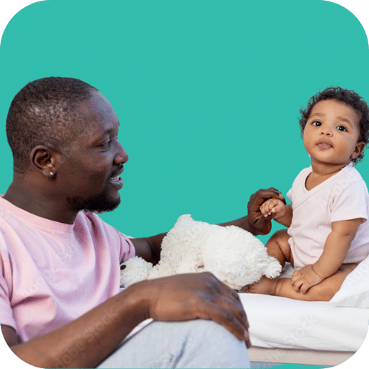 A man smiles at a baby sitting on a bed holding his hand, with a stuffed animal beside them and a teal background.