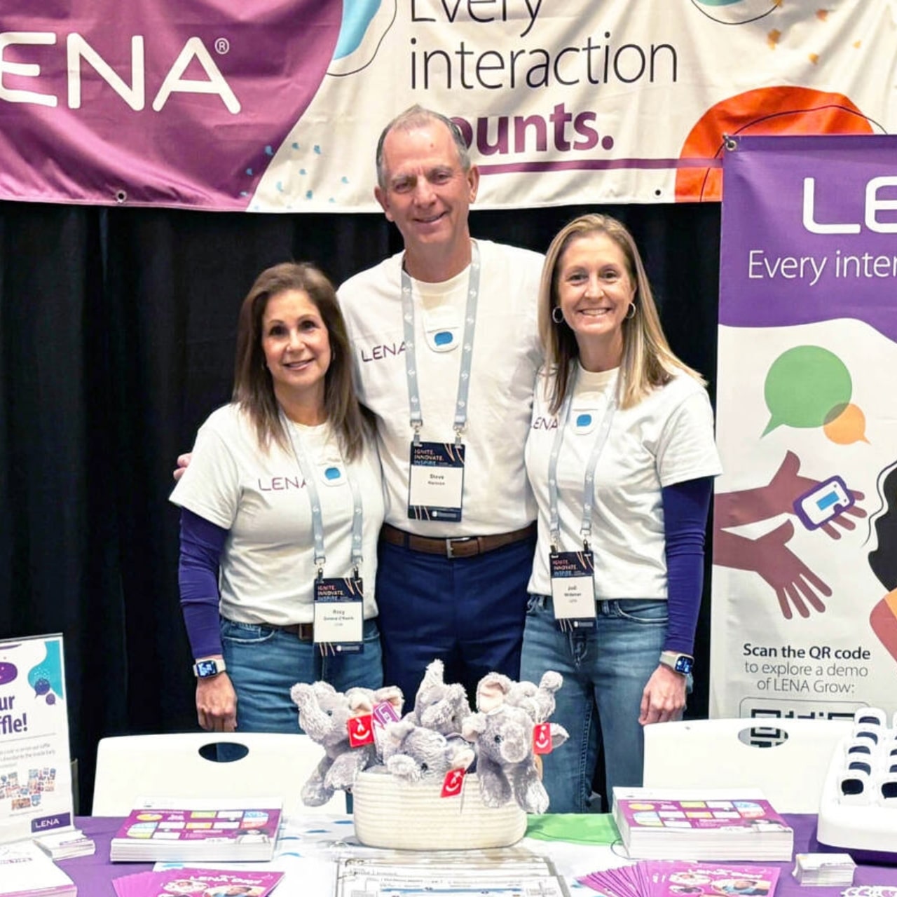 Three smiling people stand behind a booth at an event, wearing matching LENA t-shirts and name badges. The booth displays brochures and plush toys, with banners reading "Every interaction counts." in the background.