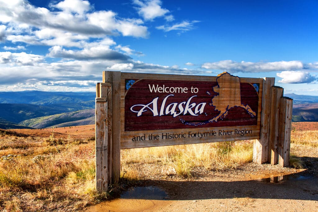 A wooden sign reads "Welcome to Alaska and the Historic Fortymile River Region," set against a scenic backdrop of rolling hills, distant mountains, and a partly cloudy blue sky.