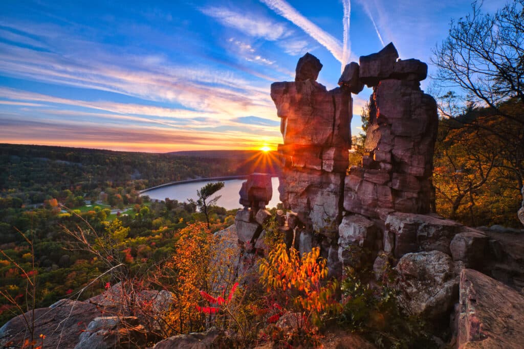 Sunset behind balanced rock formations on a cliff overlooking a lake and forest, with vibrant autumn foliage and streaks of clouds in the sky.