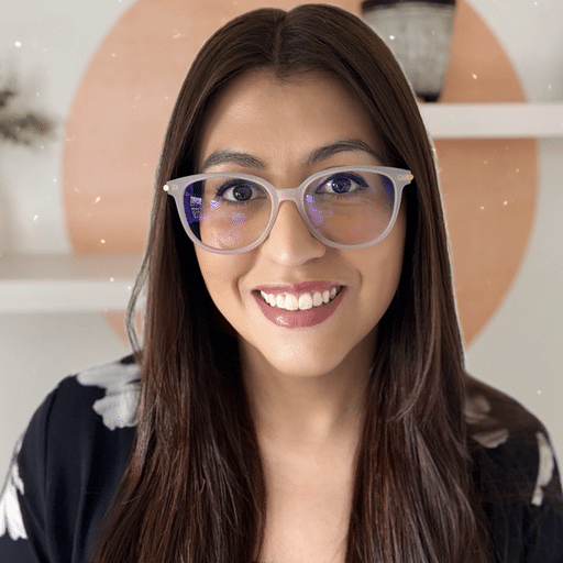 A woman with long brown hair and glasses smiles at the camera. She is wearing a black and white top and is indoors, with white shelves and a peach-colored circle on the wall behind her.