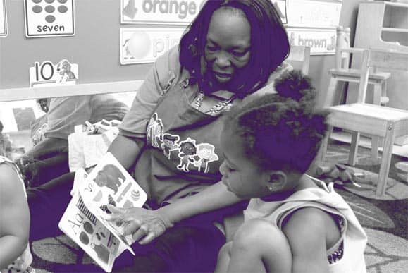 A teacher shows a picture book to a young girl in a classroom. The girl sits attentively, looking at the book while the teacher points to a page. Educational posters and a small chair are visible in the background.