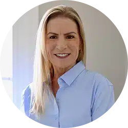 A woman with long blond hair wearing a light blue button-up shirt stands indoors, smiling at the camera. Natural light comes through a window in the background.