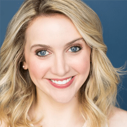 A woman with wavy blonde hair and blue eyes smiles at the camera against a solid blue background. She is wearing natural makeup and a light-colored top.