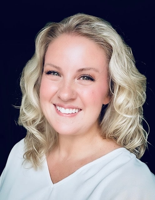 A woman with wavy blonde hair and light skin smiles at the camera. She is wearing a white top and has a nose piercing. The background is dark, making her face stand out.