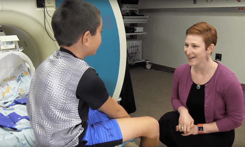 A woman with short red hair kneels and smiles while talking to a young boy sitting on the edge of a hospital MRI machine, next to a bed with colorful sheets.