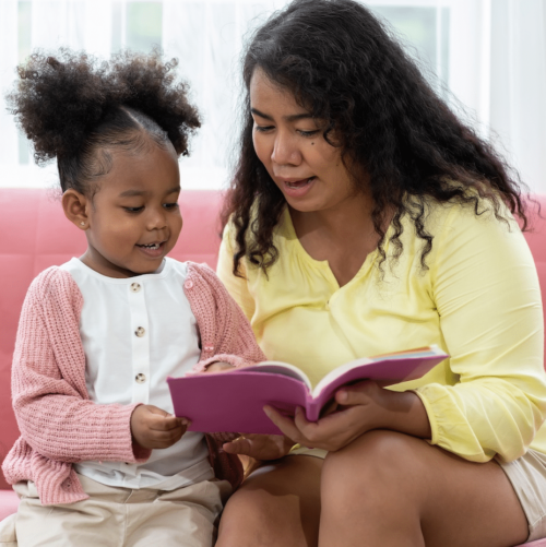 A woman and a young girl sit together on a pink couch, smiling and reading a pink book. The woman wears a yellow top, and the girl is dressed in a pink sweater and white shirt. They appear happy and engaged.