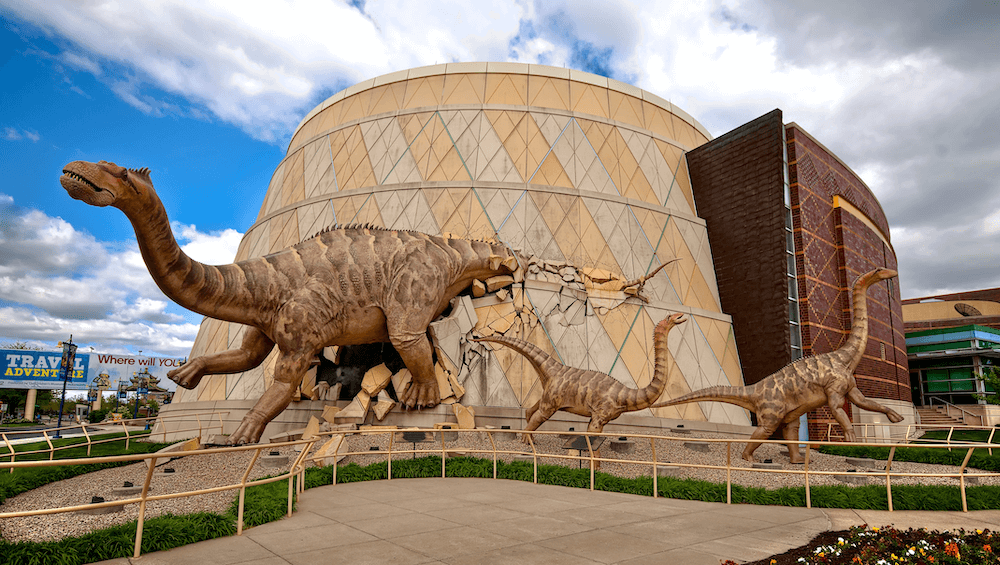 Three large dinosaur sculptures appear to burst through the wall of a modern, circular museum building, creating a dramatic scene. The sky is partly cloudy and there is greenery around the foreground.
