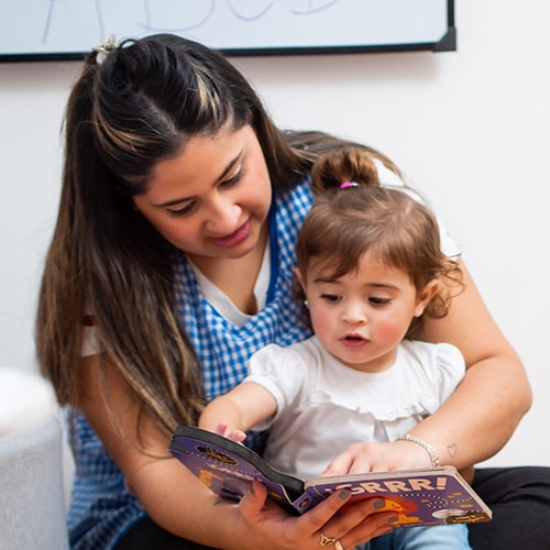 An adult woman and a young child sit together and read a colorful children’s book. The woman holds the book open while the child points at the pages, both appearing engaged and focused.