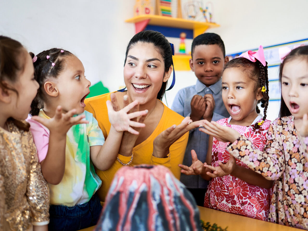A group of excited children and a smiling adult gather around a model volcano in a classroom, reacting enthusiastically, with hands raised and expressions of surprise and joy.