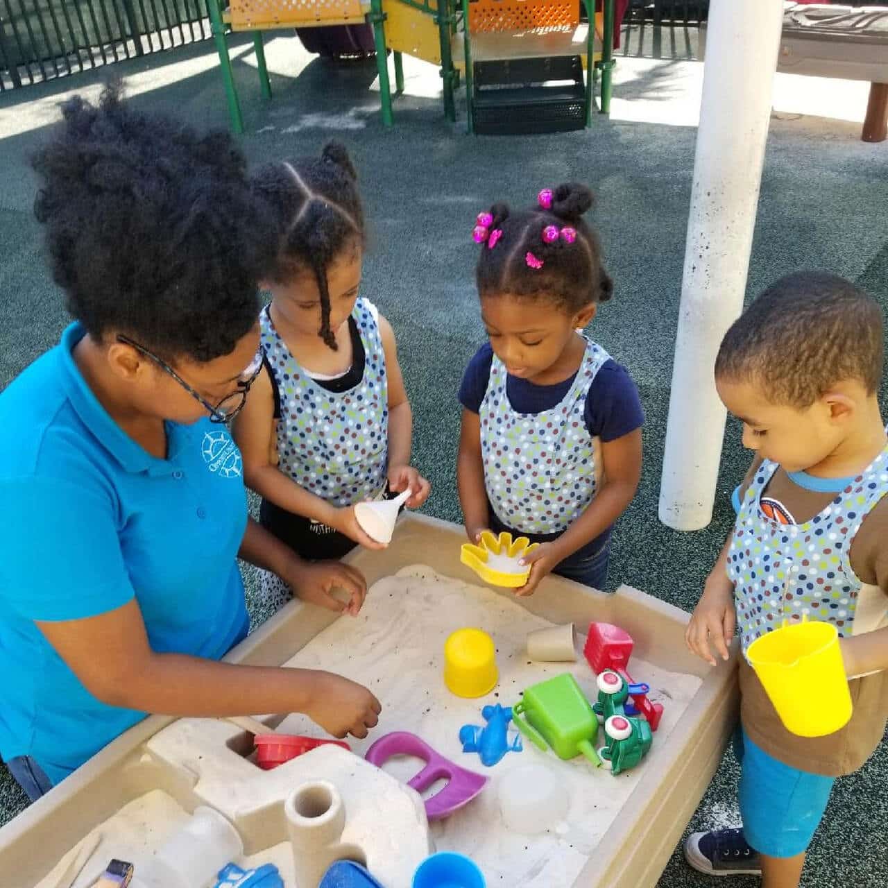 A teacher and three young children play together at a sand table outdoors, using cups, molds, and toy vehicles. The kids wear matching aprons and are engaged with the sand and toys.