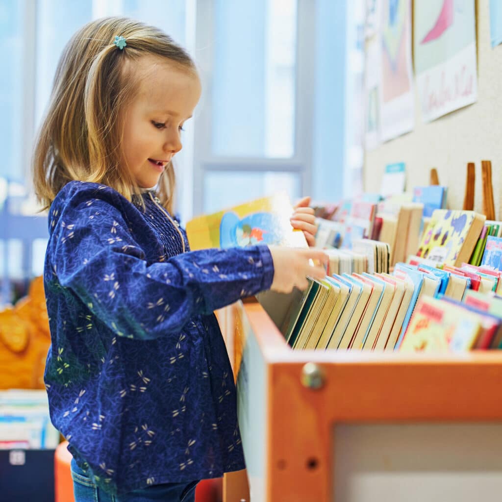 A young girl with blonde hair, wearing a blue patterned shirt, smiles as she browses through a collection of children's books in a brightly lit library or classroom.