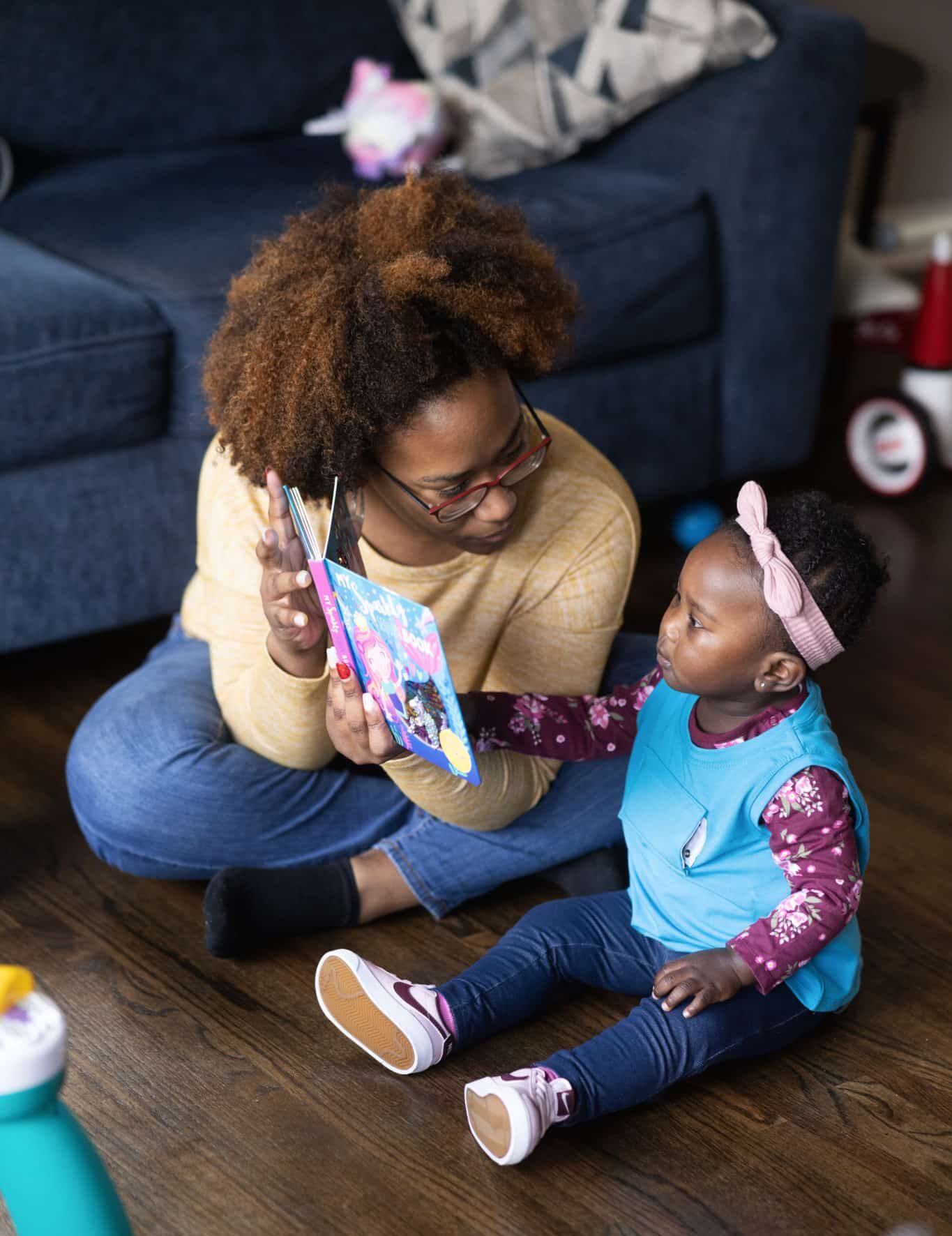 A mother sits cross-legged on the floor, holding up a colorful book and smiling at a young toddler girl beside her. The girl, wearing a headband, looks at the book and reaches toward it. They are in a cozy living room.