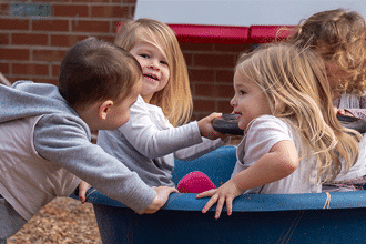Four young children are playing together outdoors, smiling and interacting in a blue playground spinner. Two girls with blonde hair face each other, while a boy and another child join in the fun. A brick wall is in the background.