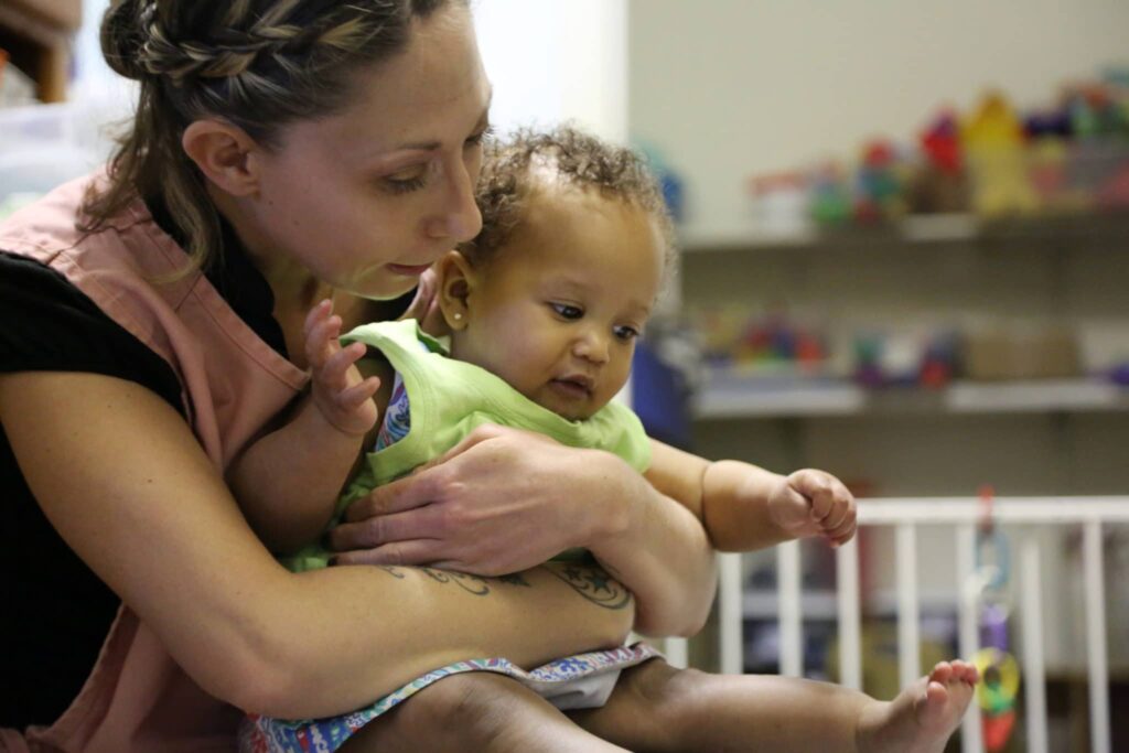 A woman gently holds a baby on her lap in a room with toys and a playpen in the background. The baby reaches forward, and the woman looks at her with care.