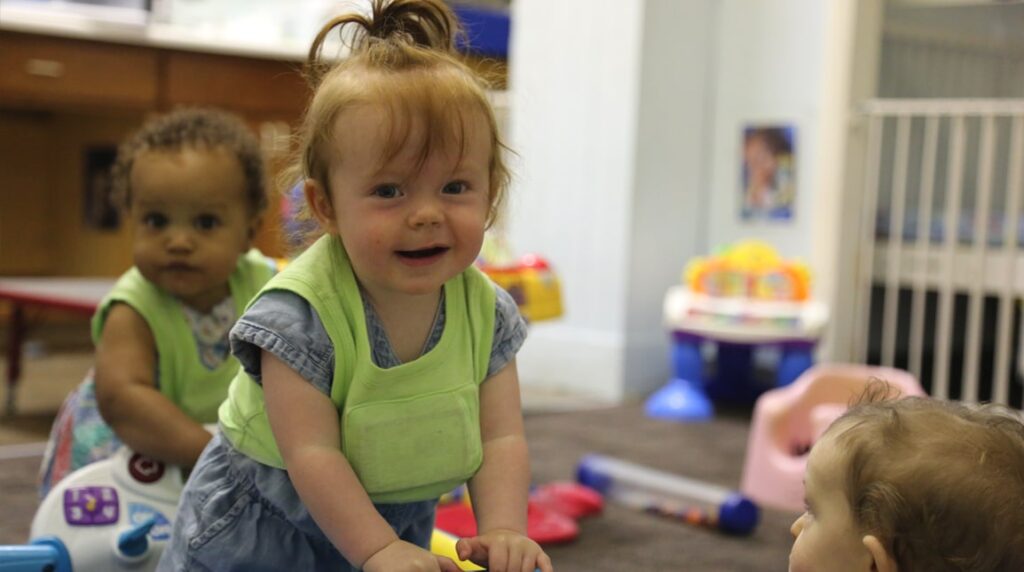 Two toddlers with green bibs play in a room filled with toys. One toddler with red hair smiles at the camera, while another child looks on. A third child is partially visible in the foreground.