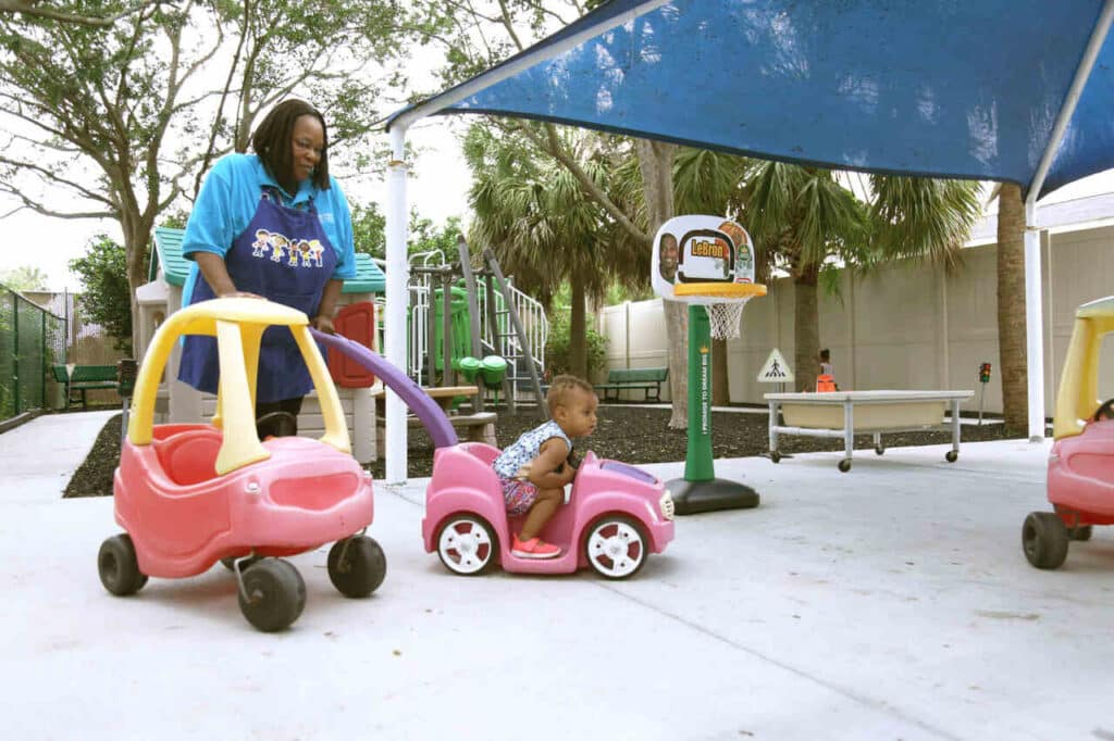A woman pushes a toddler in a pink toy car under a blue shade in an outdoor playground, with play equipment and trees in the background.