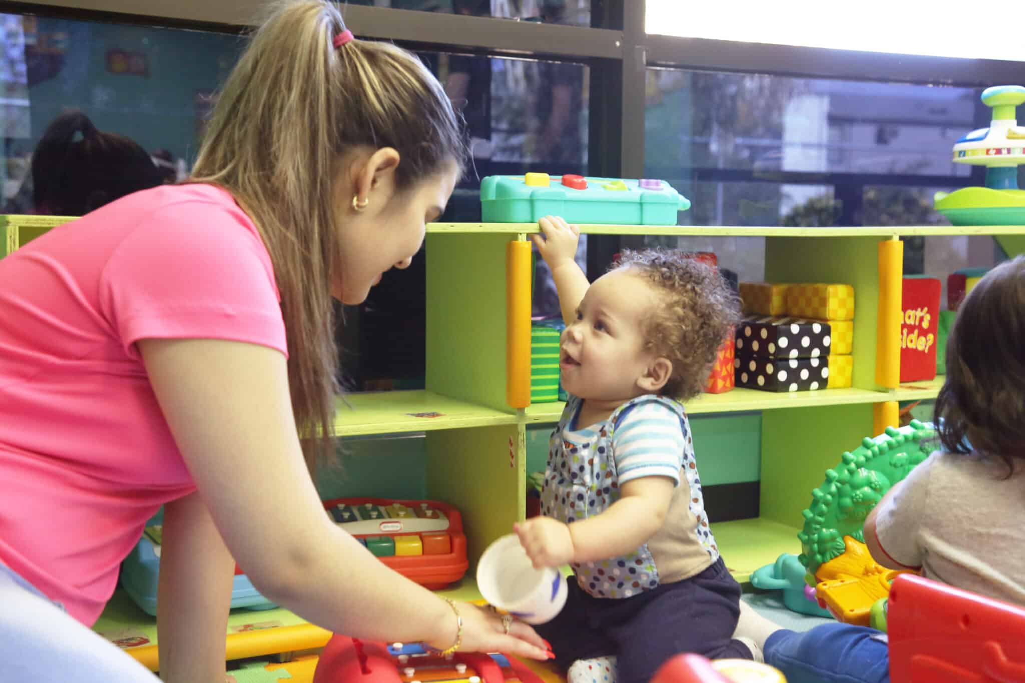 A woman in a pink shirt smiles as she interacts with a toddler holding a white cup in a colorful playroom filled with toys and shelves. Another child is partially visible on the right.