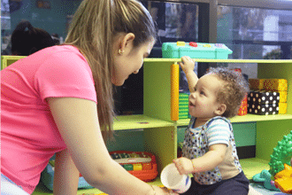 A woman smiles and interacts with a young child who is holding a cup and reaching toward her in a colorful playroom filled with toys and shelves.