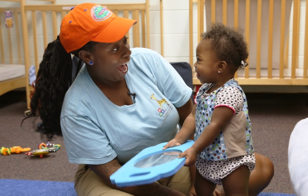 An adult and a toddler smile at each other while playing with a blue toy in a nursery, with cribs and toys visible in the background.