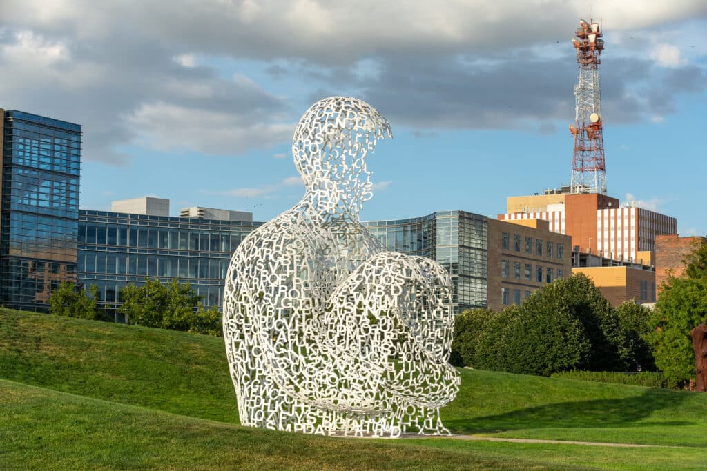 A large sculpture of a seated human figure made from interconnected white letters sits on a grassy hill, with modern office buildings and a red-and-white communication tower in the background under a partly cloudy sky.