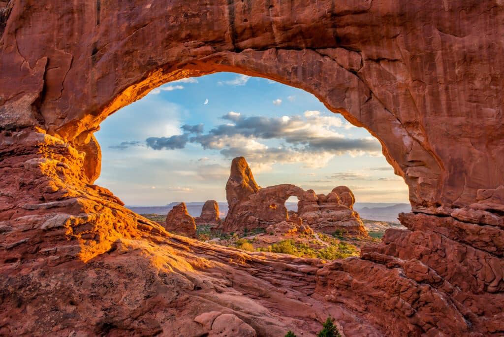 Sunlight illuminates red rock arches and formations in a desert landscape, with a partly cloudy sky and the moon visible through a large natural stone arch in the foreground.
