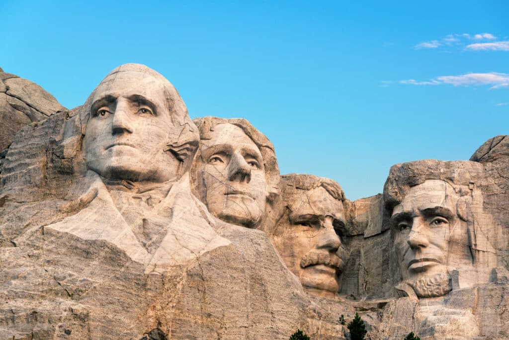 Four large stone carvings of presidential faces are sculpted into the side of a rocky mountain under a clear blue sky. The monument is Mount Rushmore.