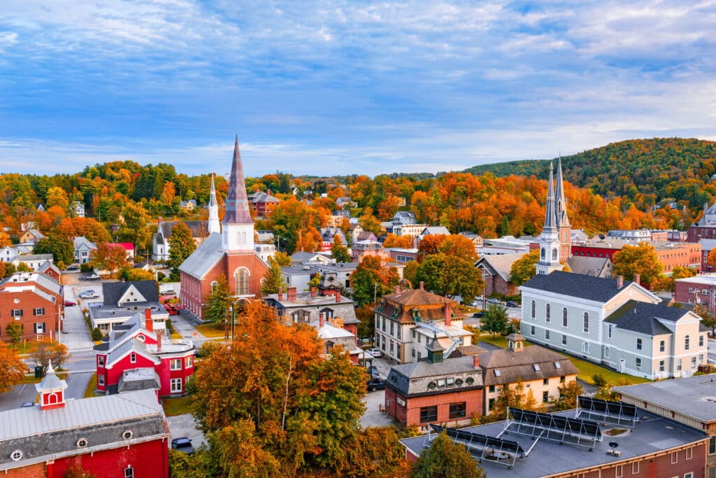 Aerial view of a small town with colorful autumn trees, historic churches with tall steeples, and red and white buildings surrounded by rolling hills under a partly cloudy sky.