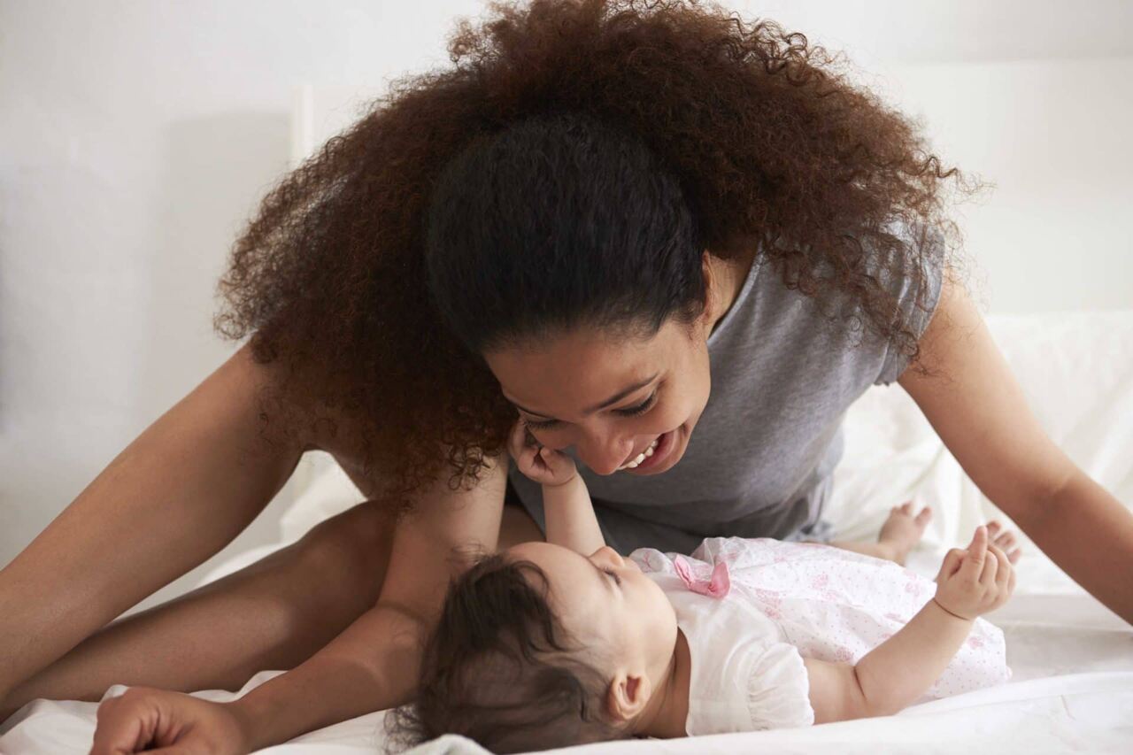 A woman with curly hair smiles lovingly at a baby while lying on a bed together. The baby reaches up towards the woman, and both appear happy and engaged with each other in a bright, cozy setting.