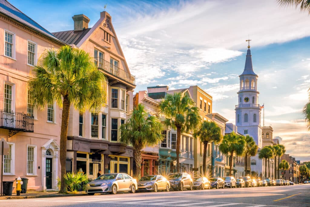 Colorful historic buildings and palm trees line a sunlit street with parked cars. A white church steeple rises in the background under a partly cloudy sky.