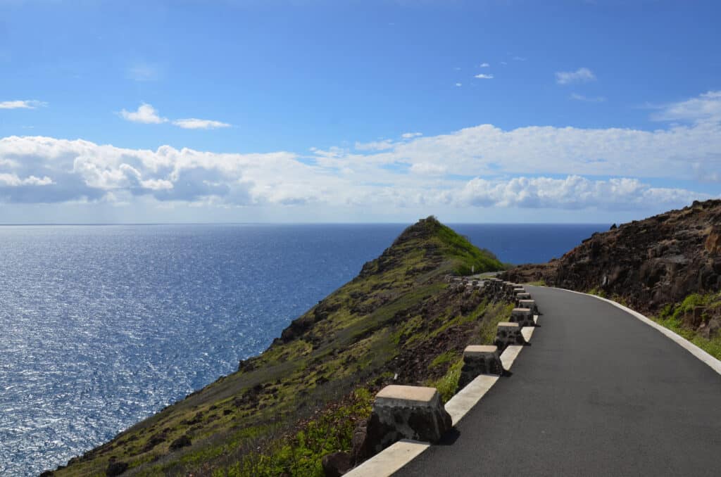 A paved road curves along a rocky hillside overlooking a vast, sparkling blue ocean under a bright sky with scattered clouds.