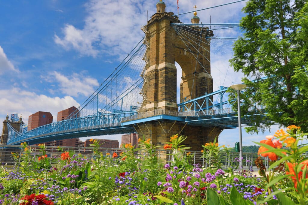 A blue suspension bridge spans over a colorful flower garden with red, orange, and purple blooms, set against a backdrop of city buildings and a partly cloudy sky.
