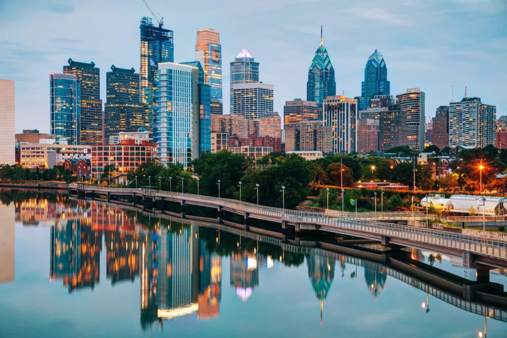 Philadelphia skyline at dusk, with modern skyscrapers and buildings reflected in the calm water of the Schuylkill River, and a pedestrian path running alongside the river.
