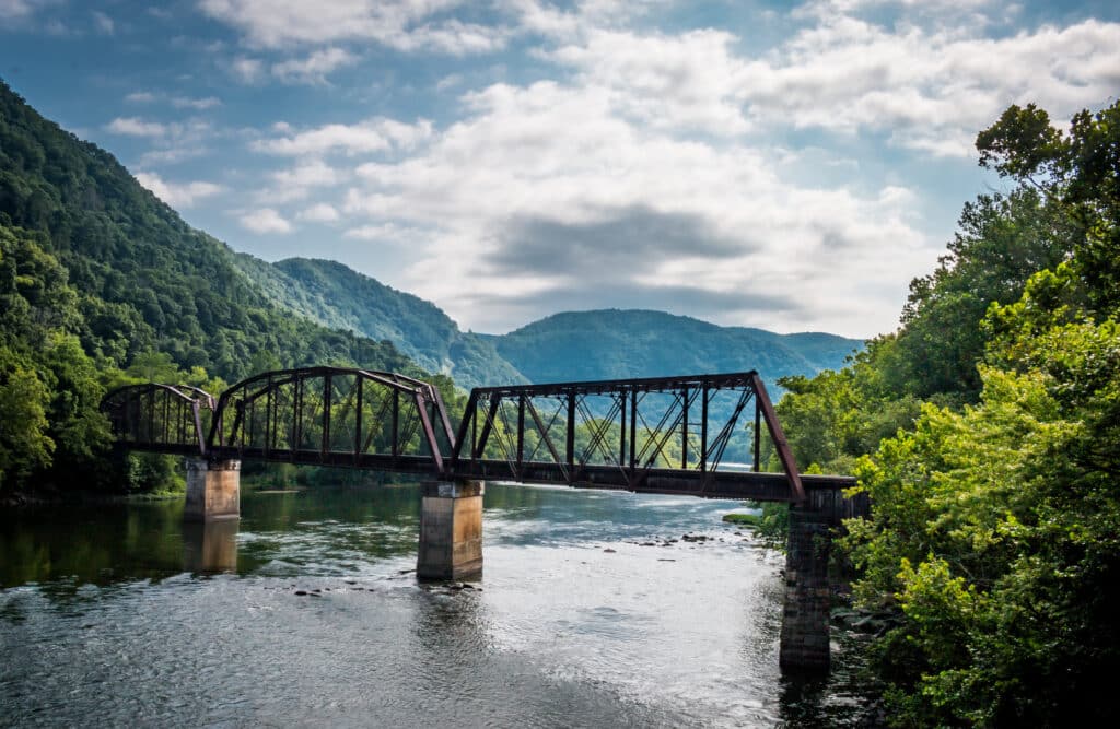 A steel railway bridge with trusses spans a calm river, surrounded by lush green trees and rolling hills under a partly cloudy blue sky.