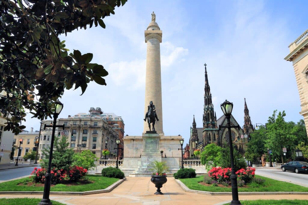 A tall monument with a statue at its base stands in a city square surrounded by colorful flowers, historic buildings, and a church with a tall spire under a partly cloudy sky.