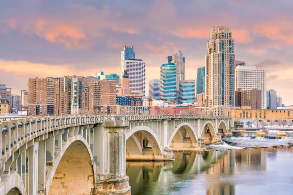 A city skyline at sunset with modern and historic buildings, a large arched bridge crossing a river in the foreground, and colorful clouds in the sky.